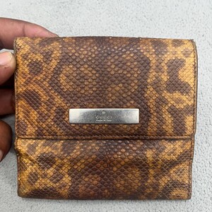 Op de afbeelding: Een vierkante portemonnee met een bruine en beige textuur in een slangenleerpatroon. Op de voorflap bevindt zich in het midden een rechthoekig, zilverkleurig metalen plaatje, gegraveerd met de tekst GUCCI en daaronder Made in Italy.