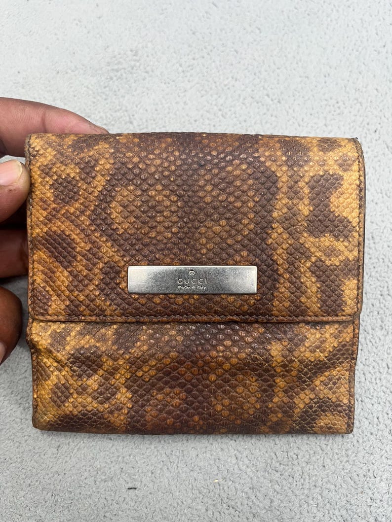 May include: A square-shaped wallet featuring a brown and tan snakeskin-patterned texture. A rectangular silver-toned metal plaque is centered on the front flap, engraved with the text GUCCI and the words Made in Italy below the brand name.