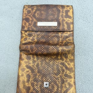 May include: A rectangular wallet featuring a brown and yellow snakeskin pattern. The wallet has a fold-over flap with a silver-toned metal bar engraved with the word GUCCI. A small silver-toned metal snap button is visible on the lower front section.