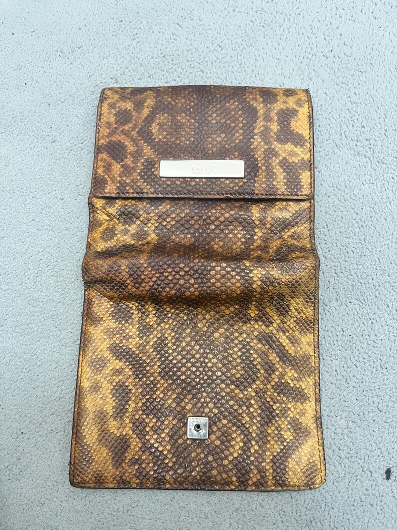 May include: A rectangular wallet featuring a brown and yellow snakeskin pattern. The wallet has a fold-over flap with a silver-toned metal bar engraved with the word GUCCI. A small silver-toned metal snap button is visible on the lower front section.