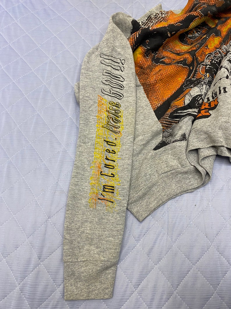 May include: A heather gray long-sleeve sweatshirt featuring a large, multi-colored graphic print on the chest and the text "I'm Cured. Praise God!!" printed vertically in black and yellow along the length of the left sleeve.