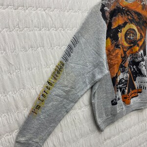 May include: A heather gray long-sleeve sweatshirt featuring a large orange and black graphic print on the front and the text "I'm cured! Praise God!!" printed in black and yellow along the sleeve. The graphic includes the text "Clockwork Orange".
