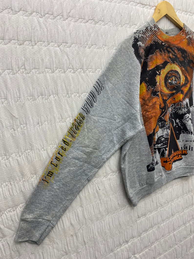 May include: A heather gray long-sleeve sweatshirt featuring a large orange and black graphic print on the front and the text "I'm cured! Praise God!!" printed in black and yellow along the sleeve. The graphic includes the text "Clockwork Orange".