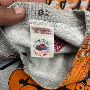 May include: A grey sweatshirt with a Fruit of the Loom tag reading Ladies XL, 50% Cotton, 50% Polyester, Made in U.S.A. The collar features the number 82 printed in black. The fabric has a textured grey interior and orange graphic print details.