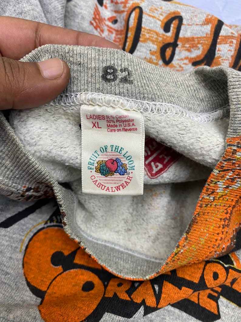 May include: A grey sweatshirt with a Fruit of the Loom tag reading Ladies XL, 50% Cotton, 50% Polyester, Made in U.S.A. The collar features the number 82 printed in black. The fabric has a textured grey interior and orange graphic print details.