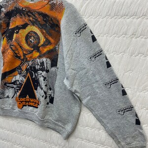 May include: A heather gray crewneck sweatshirt featuring a large, vibrant orange and black graphic print of an eye and film-related imagery on the front. The text "CLOCKWORK ORANGE" is printed on the front and repeatedly down the right sleeve.