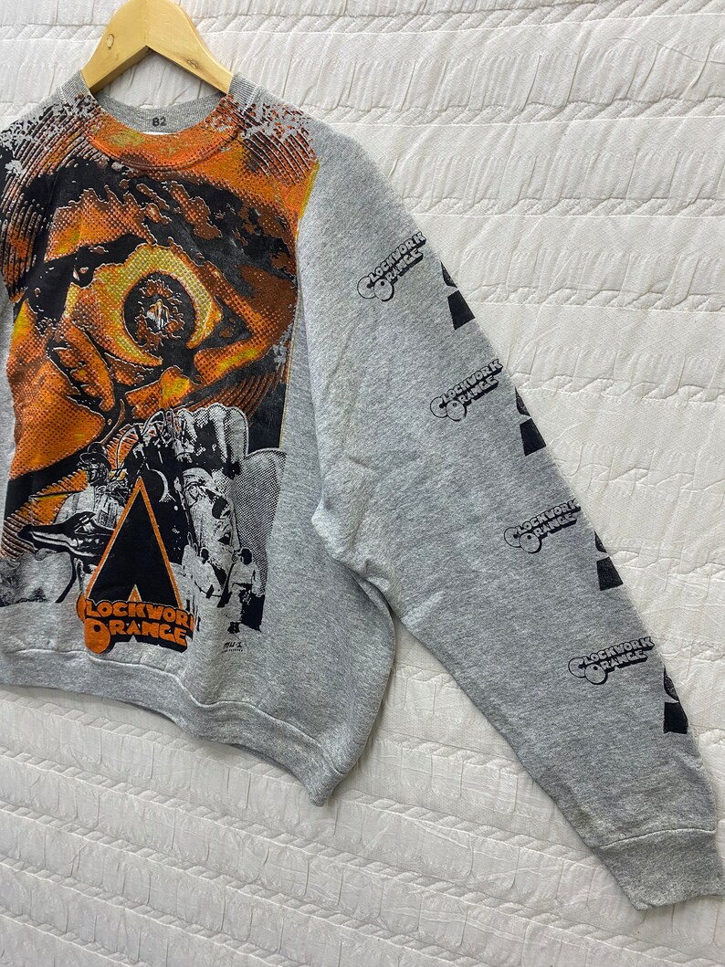 May include: A heather gray crewneck sweatshirt featuring a large, vibrant orange and black graphic print of an eye and film-related imagery on the front. The text "CLOCKWORK ORANGE" is printed on the front and repeatedly down the right sleeve.