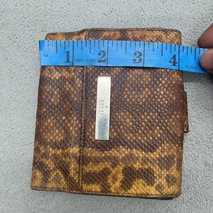 May include: A brown and tan snakeskin-patterned wallet featuring a rectangular silver-toned metal plaque engraved with the Gucci logo. A blue measuring tape is placed across the top, indicating a width of approximately 4 inches.