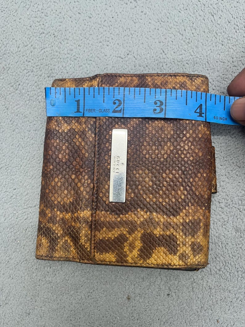 May include: A brown and tan snakeskin-patterned wallet featuring a rectangular silver-toned metal plaque engraved with the Gucci logo. A blue measuring tape is placed across the top, indicating a width of approximately 4 inches.