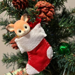 Peut inclure: Ornement de Noël sur un sapin. Il présente une chaussette rouge en feutre avec une bordure blanche et une petite tête de cerf en peluche. La chaussette est cousue avec du fil blanc et est suspendue parmi des pommes de pin et des branches vertes.
