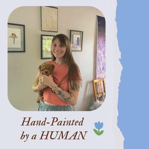 Może przedstawiać: Uśmiechnięta kobieta trzyma małego, brązowego psa. Obraz zawiera oprawione dzieła sztuki na ścianie i tekst "Hand-Painted by a HUMAN" z kwiatową grafiką.