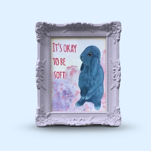 May include: A framed print with a watercolour background and a blue rabbit. The text "It's okay to be soft" is written in pink.