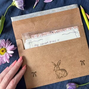 Może przedstawiać: Brązowa koperta z papieru z ilustracją kr&oacute;lika i motywami kokardek, zawierająca kartkę z napisem "my newest friend". Scena obejmuje fioletowe kwiaty i kredki na niebieskim tle.