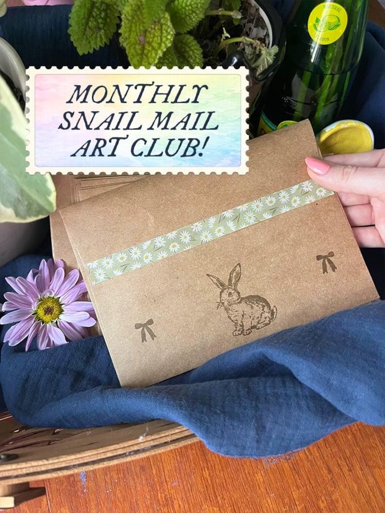 May include: Two brown paper envelopes with daisy-patterned tape and a rabbit illustration. The envelopes are labeled "MONTHLY SNAIL MAIL ART CLUB!" in a colorful stamp design. A purple flower and a blue cloth are in the background.