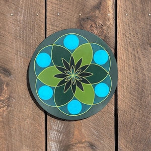 May include: A round, decorative wooden piece with a geometric floral design. The design features a dark green background with a central flower shape in black and green, surrounded by blue circles and yellow outlines.