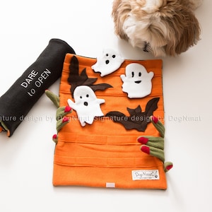 Könnte beinhalten: Ein schwarzes und oranges Halloween-Hundespielzeug mit dem Text "Dare to Open" und einem weißen Geist mit einem lächelnden Gesicht. Das Spielzeug liegt auf einer weißen Oberfläche, auf der ein kleiner weißer Hund daran schnuppert.