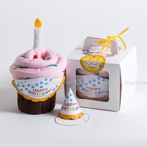 Może przedstawiać: Różowo-brązowy koc dla psa w kształcie babeczki z żółtą świeczką na górze, pasujący kapelusz imprezowy i przezroczyste pudełko z okienkiem pokazującym koc w środku. Pudełko ma żółtą wstążkę i etykietę z napisem "Dog Nmat". Na kocu i kapeluszu nadrukowany jest napis "Happy Birthday!"