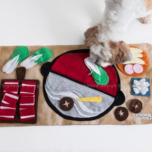 Könnte beinhalten: Eine braune Filz-Spielmatte für Hunde mit verschiedenen Filzformen, darunter ein roter und weißer Topf, ein Schneidebrett mit rotem und weißem Fleisch, grünem Gemüse und einem braunen Pilz. Der Text "DogNmat Signature design by DogNmat" ist auf der Matte aufgedruckt.