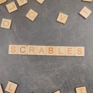 Puede incluir: Fichas de Scrabble de madera que deletrean "SCRABLES" sobre una superficie gris. Fichas individuales con letras están dispersas alrededor de la palabra. Las fichas son de color marrón claro con letras grabadas.
