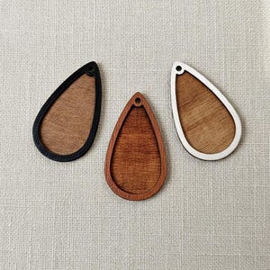 Puede incluir: Tres colgantes de madera en forma de lágrima. Uno tiene un borde negro, otro un borde de madera natural y el tercero un borde blanco. Cada colgante tiene un pequeño agujero en la parte superior para colgar.