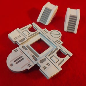 May include: White and gray miniature building components on a red surface. The set includes a large, multi-faceted piece with a square opening and two smaller stairwell structures. The pieces have gray detailing.