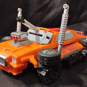 May include: Orange toy vehicle with black treads and wheels. Features a silver engine and exhaust pipes. The toy has a gray roof and a red and orange design on the top. The vehicle is a toy.