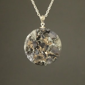 May include: A silver necklace with a circular pendant. The pendant has a distinctive abstract design, incorporating black, grey, and gold elements within a clear resin. The chain is a fine silver link.