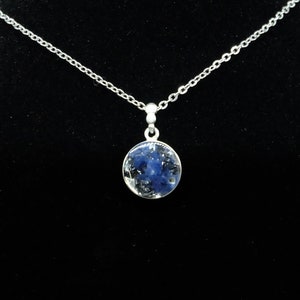 Op de afbeelding: Een zilveren ketting met een ronde hanger met een blauwe steen ingekapseld in helder hars.
