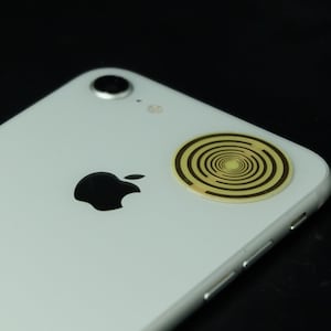 Könnte beinhalten: Ein weißes iPhone mit einem goldenen runden Aufkleber auf der Rückseite. Der Aufkleber hat ein Spiraldesign.