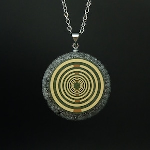 May include: A silver-toned necklace with a circular pendant featuring a concentric spiral design in shades of green, gold, and brown. The pendant is set against a textured, dark gray border, suspended from a delicate chain.