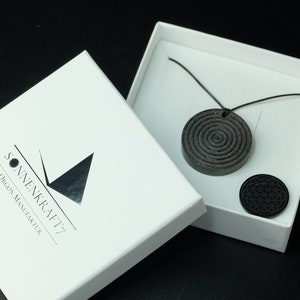 Könnte beinhalten: Eine schwarze Halskette mit einem spiralförmigen Anhänger und einem kleineren schwarzen Anhänger mit einem geometrischen Muster. Die Halskette befindet sich in einer weißen Schachtel mit dem aufgedruckten Text "SONNENKRAFT7 ORGON MASTERTUR".