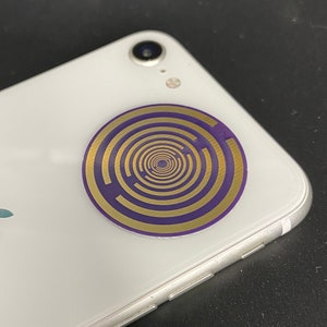 Puede incluir: Un smartphone blanco con un diseño circular dorado y morado en la parte trasera. El diseño presenta un patrón en espiral. El teléfono tiene una lente de cámara y el logotipo de Apple.