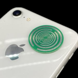 Könnte beinhalten: Ein weißes Smartphone mit einem grünen und silbernen kreisförmigen Design auf der Rückseite. Das Design weist ein Spiralmuster auf. Das Telefon hat ein silbernes Apple-Logo und eine Kameralinse.