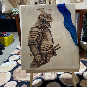 May include: A wood burned image of a samurai warrior in full armor, with a blue resin river flowing down the side of the wood panel. The panel is on a wooden easel.