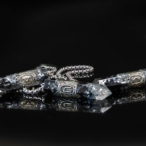 Puede incluir: Tres collares con colgantes de cristal y cadenas plateadas. Los cristales son transparentes con inclusiones oscuras. Los collares se exhiben sobre una superficie negra reflectante.