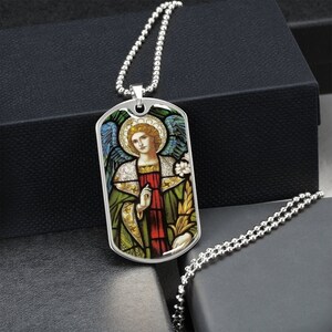 Puede incluir: Un collar de cadena de plata con un colgante de placa de identificaci&oacute;n de plata que presenta una imagen de vitral de un &aacute;ngel con alas, una t&uacute;nica verde y una faja roja.