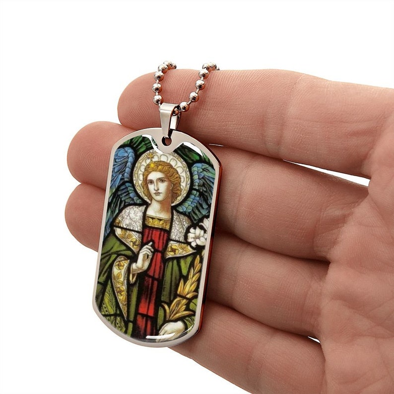 Puede incluir: Un collar con una placa de identificaci&oacute;n de perro de plata con una imagen de vitral de un &aacute;ngel con alas, un halo y una t&uacute;nica verde y roja.