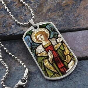 Puede incluir: Un collar de cadena plateada con un colgante de placa de identificaci&oacute;n de acero inoxidable con una imagen de vitral de un &aacute;ngel con alas, un halo y una t&uacute;nica verde y roja.