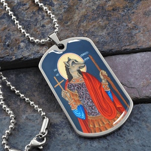 May include: A silver chain necklace with a stainless steel dog tag pendant featuring a colorful icon of Saint Christopher. The icon depicts Saint Christopher as a wolf-like figure holding a staff and a cross. The text "SAN NZ" and "CHRISSOCO" are visible on the dog tag.