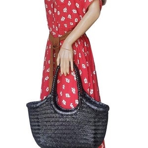 May include: A black woven tote bag with a curved shape and braided handles. The bag is held by a mannequin wearing a red dress with white floral patterns and a brown belt.