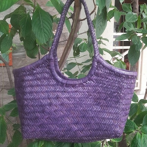 May include: A woven purple tote bag with braided handles. The bag has a rectangular shape with a wide opening and a textured, basket-like weave. The bag is hanging from a tree branch, surrounded by green leaves.