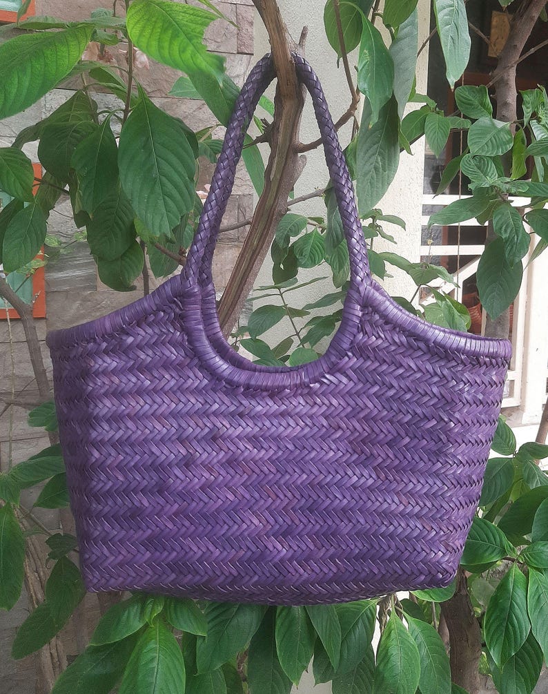 May include: A woven purple tote bag with braided handles. The bag has a rectangular shape with a wide opening and a textured, basket-like weave. The bag is hanging from a tree branch, surrounded by green leaves.