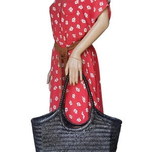 May include: A black woven tote bag with a braided handle. The bag is held by a mannequin wearing a red dress with white floral patterns and a brown belt. The bag's dimensions are approximately 18 inches wide and 12 inches tall.