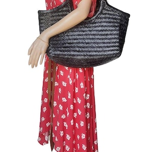 May include: A black woven tote bag with a rectangular shape is held by a mannequin wearing a red floral dress. The bag has a wide opening and a woven pattern. The dress has a brown belt.