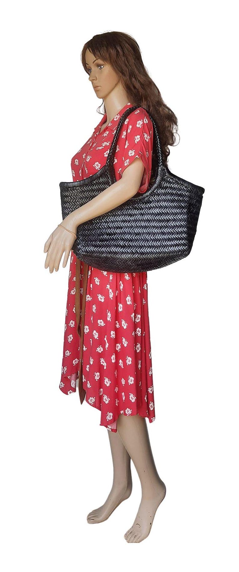 May include: A black woven tote bag with a rectangular shape is held by a mannequin wearing a red floral dress. The bag has a wide opening and a woven pattern. The dress has a brown belt.