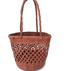 May include: A woven brown tote bag with a braided handle. The bag has a round base and a lattice-like pattern on the sides. The handles are also braided. The bag is made of a natural material.
