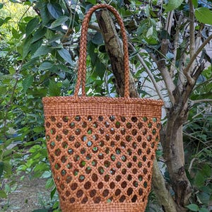 May include: A woven brown tote bag with a lattice design. The bag has a braided handle and is hanging from a tree branch. The bag is made of a natural material and has a spacious interior.