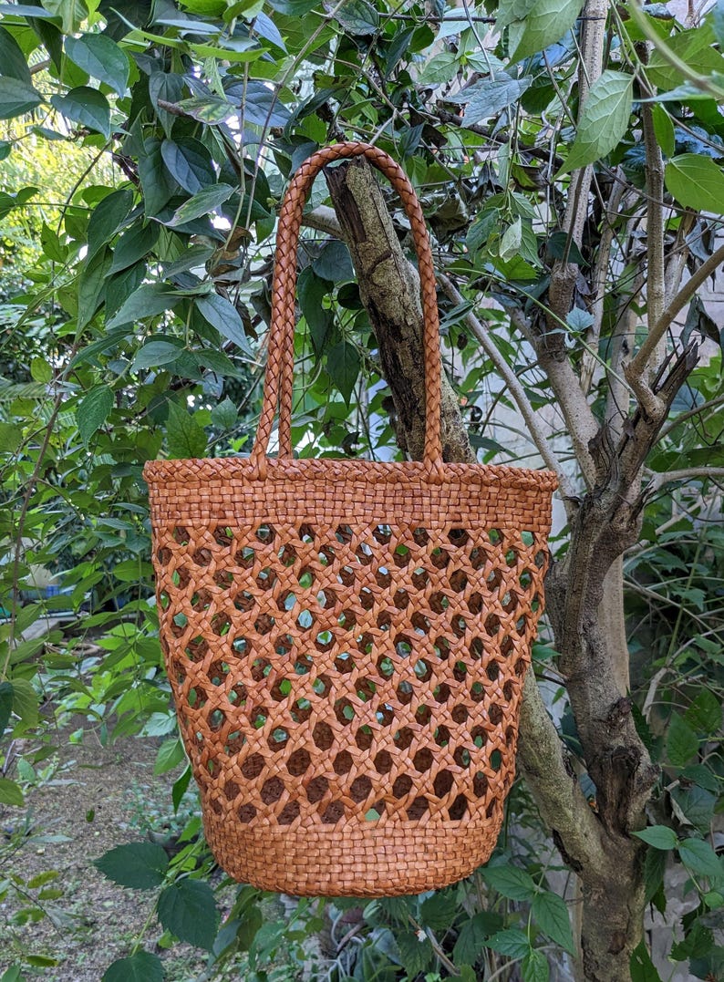 May include: A woven brown tote bag with a lattice design. The bag has a braided handle and is hanging from a tree branch. The bag is made of a natural material and has a spacious interior.