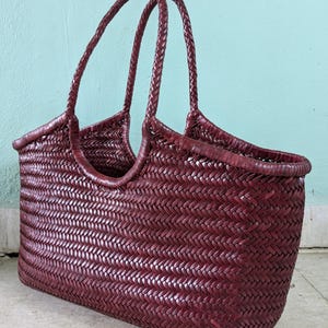 May include: A burgundy woven tote bag with braided handles. The bag features a rectangular shape with a wide opening and a textured, basket-like weave. The handles are arched and attached to the sides of the bag.