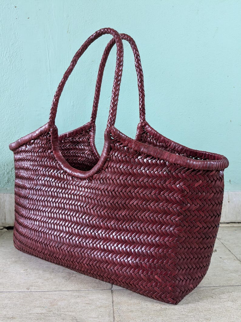 May include: A burgundy woven tote bag with braided handles. The bag features a rectangular shape with a wide opening and a textured, basket-like weave. The handles are arched and attached to the sides of the bag.